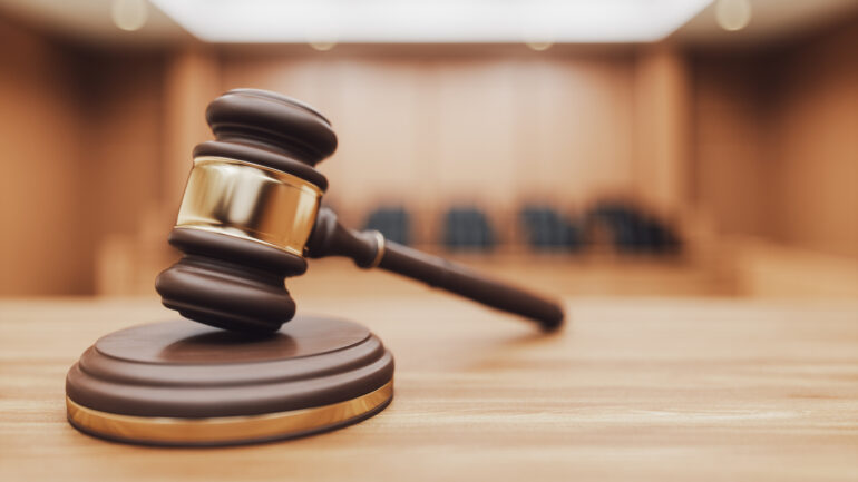 A wooden judge's gavel and sounding block on a desk with a blurred courtroom in the background. Focus on themes of law, authority, and verdicts.