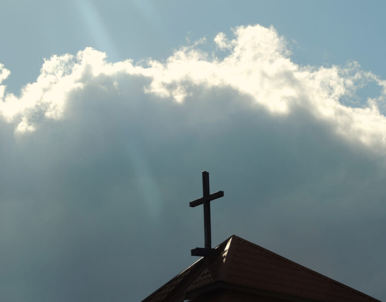 simple dark cross on top of protestant church roof against stormy beautiful sky, concept of faith in god against contrast of dark cloud against shining sky with cross in center
