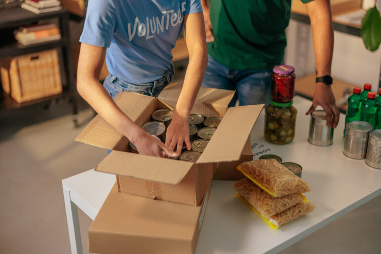Two volunteers arranging various food donations in cardboard boxes for distribution in a local charity food bank
