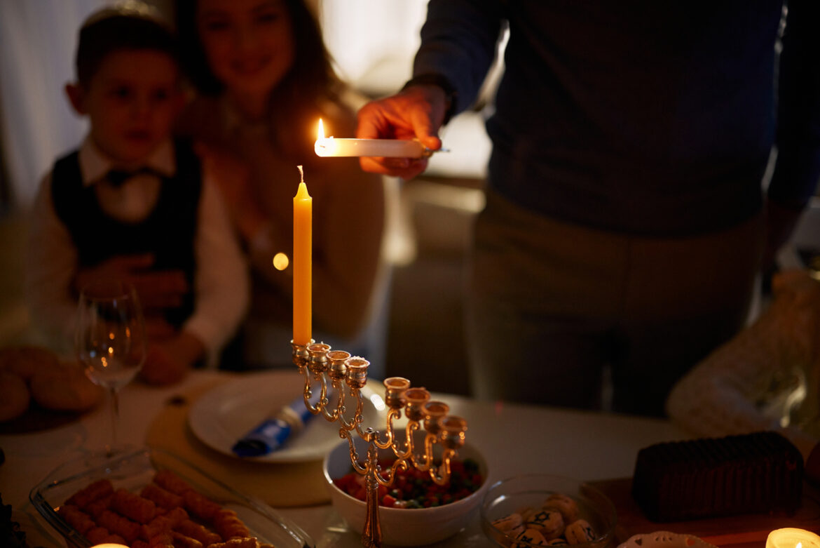 Close up of unrecognizable man lightning candles on Menorah before Hanukkah meal with his family in dining room.