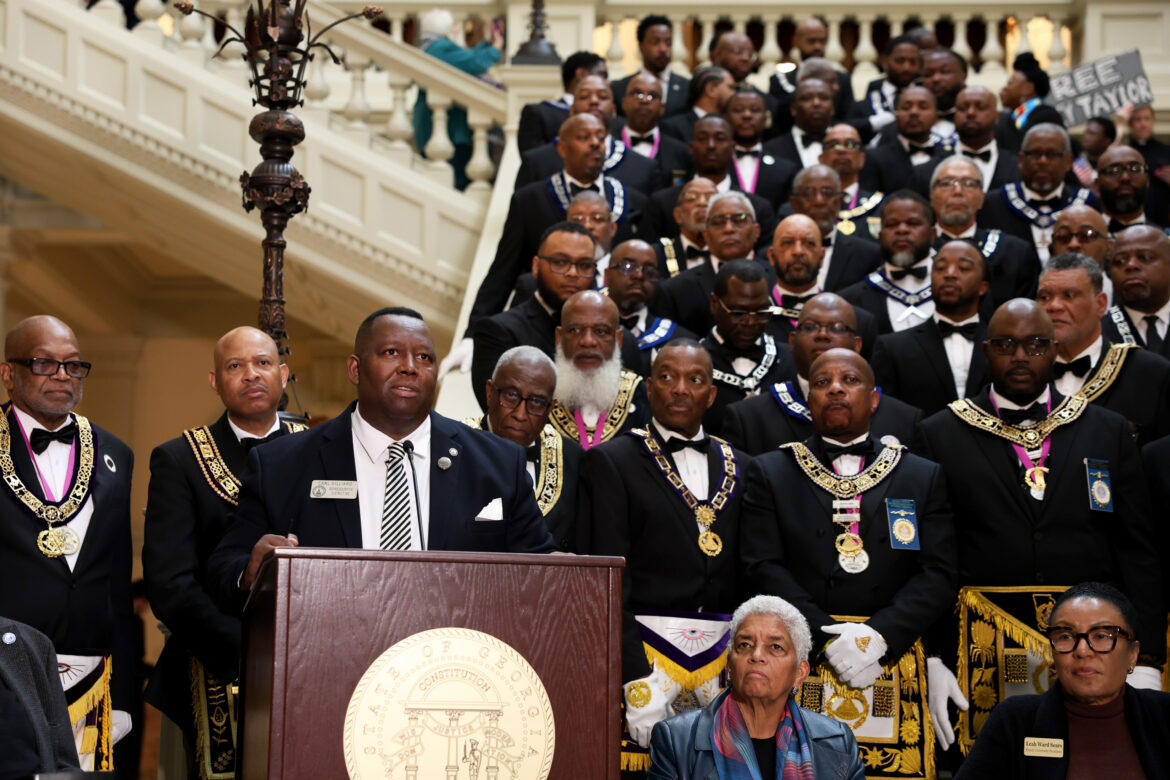A medium shot of State Rep. Carl Gilliard giving a speech at the bottom of the South Wing steps at the Georgia State Capitol. Several supporters stand behind Gilliard.