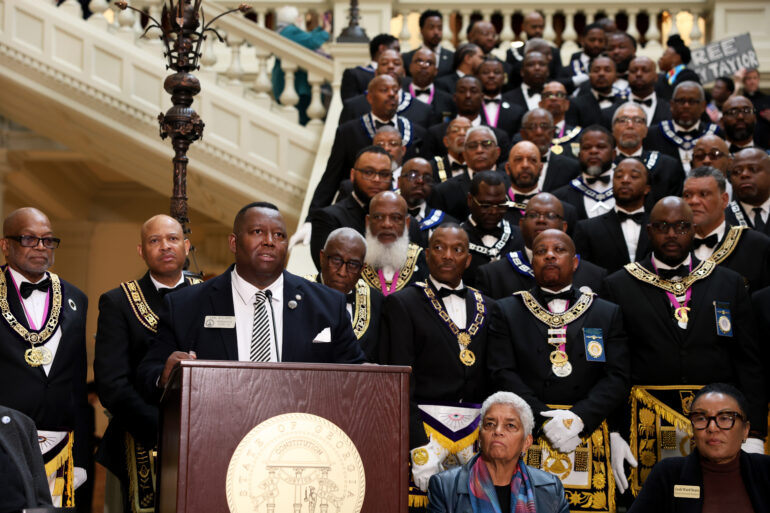 A medium shot of State Rep. Carl Gilliard giving a speech at the bottom of the South Wing steps at the Georgia State Capitol. Several supporters stand behind Gilliard.