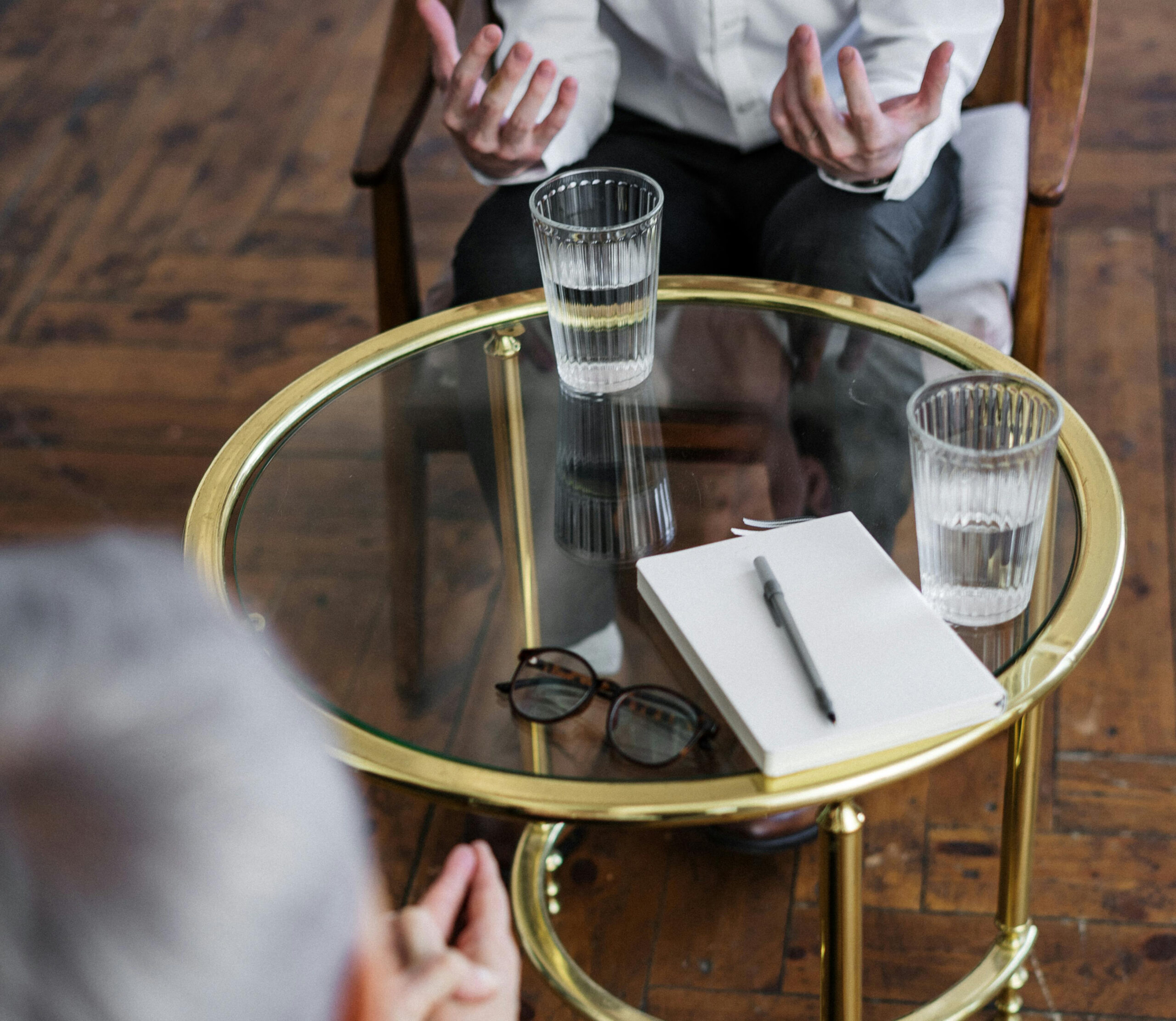 A close up photo of the back of a therapist's head as they are talking to a patient who is holding their hands held out. The table has two glasses of water and a notepad with a pen on it.